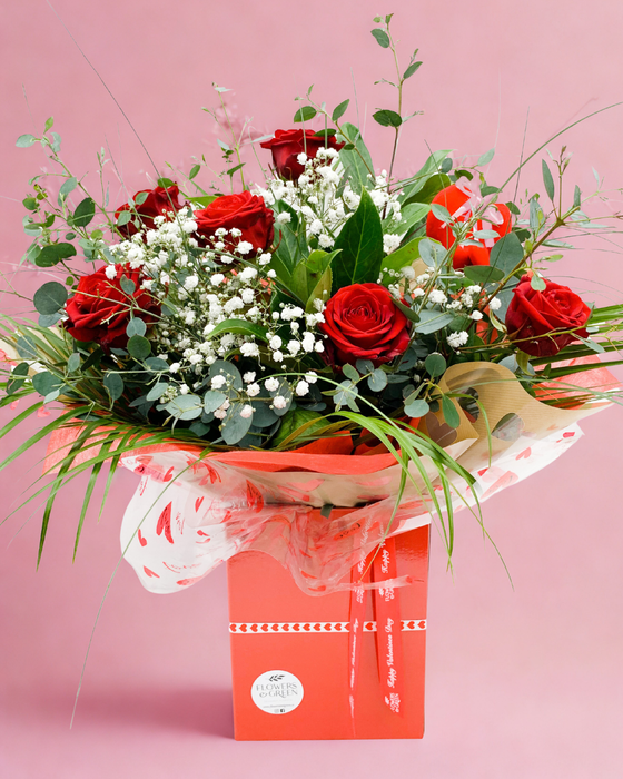 A bouquet of red roses with baby's breath and green foliage, presented in a red Valentine's gift box with water.
